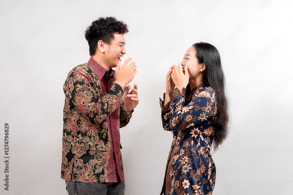 A young couple, dressed in vibrant Indonesian batik shirts, laugh together against a simple gray background. Their joyful expressions and stylish attire showcase Indonesian cultural fashion.
