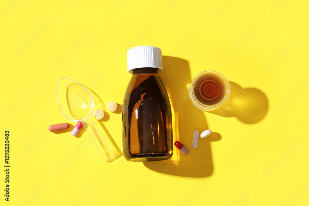 Bottle of cough syrup with measuring cup, spoon and different pills on yellow background Bottle of cough syrup with measuring cup, spoon and different pills on yellow background