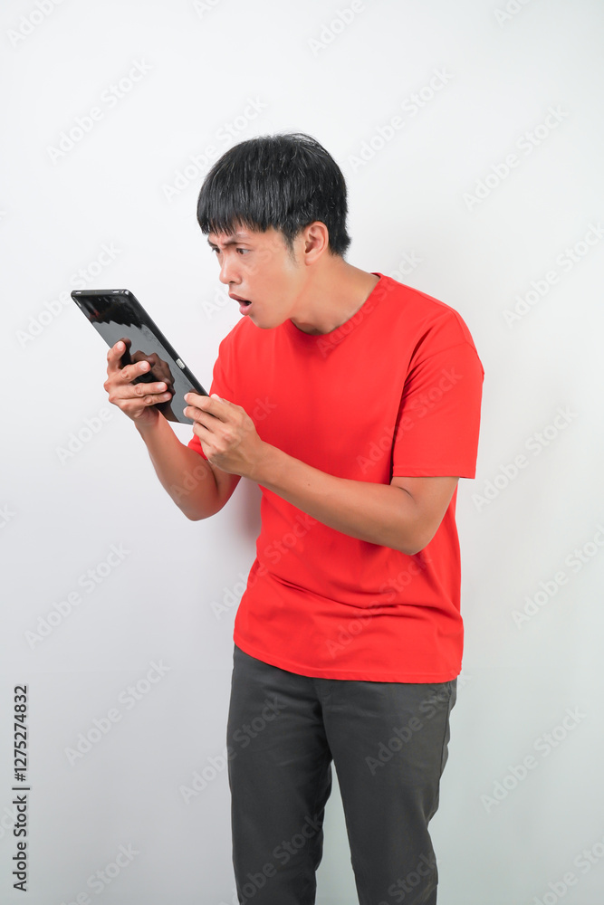 Amazed young asian man wearing red shirt is holding a tablet screen, isolated over white background. Amazed young asian man wearing red shirt is holding a tablet screen, isolated over white background.