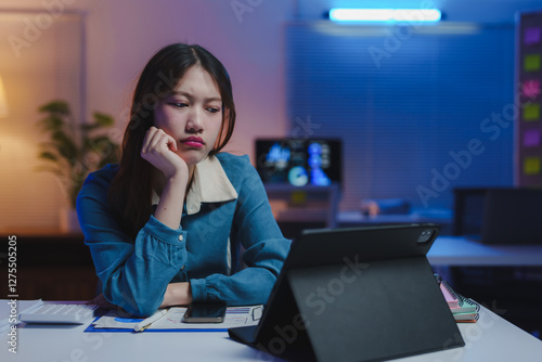 Young asian businesswoman with a bored expression resting her face on her hand while looking at a tablet in a dimly lit office at night, surrounded by paperwork and technology