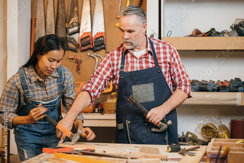 Obraz na plátně Senior carpenter teaches a woman woodworking techniques, guiding her as she uses a hammer on a wood project
