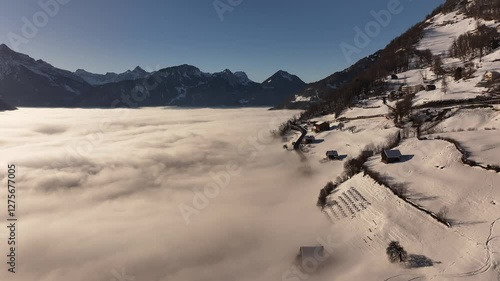 Aerial view of Amden, Switzerland, revealing a mesmerizing winter landscape where snow-covered slopes meet a dense sea of clouds. The contrast between the crisp alpine peaks and the misty valley below