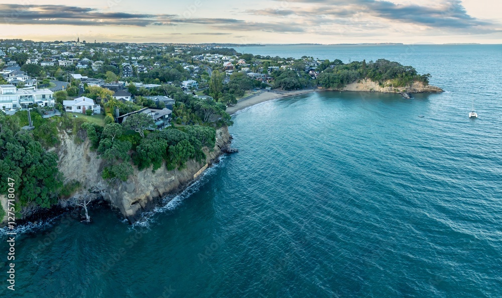 Fototapeta premium Coastal town with a secluded beach, a catamaran in the bay. Tranquil scene from above. CASTOR BAY, AUCKLAND, NEW ZEALAND