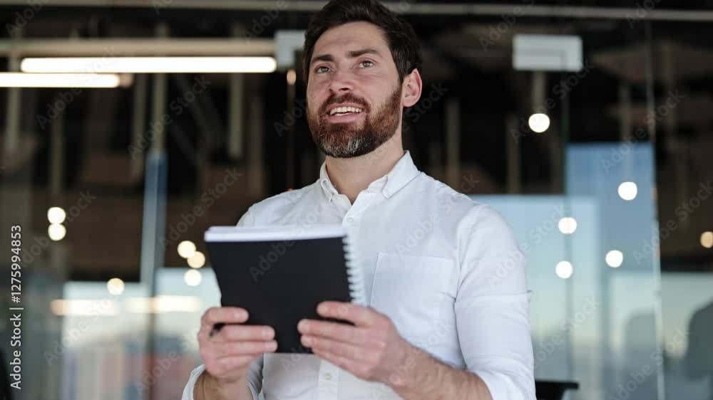 Confident businessman takes notes in notebook, looking around modern office. Bearded man in white shirt stands in modern office. Confident businessman takes notes in notebook, looking around modern office. Bearded man in white shirt stands in modern office.