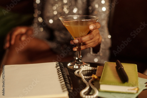 Person holding cocktail glass in elegant setting with pearl necklace and notebook on table. Sparkling dress and soft lighting creating sophisticated atmosphere photo