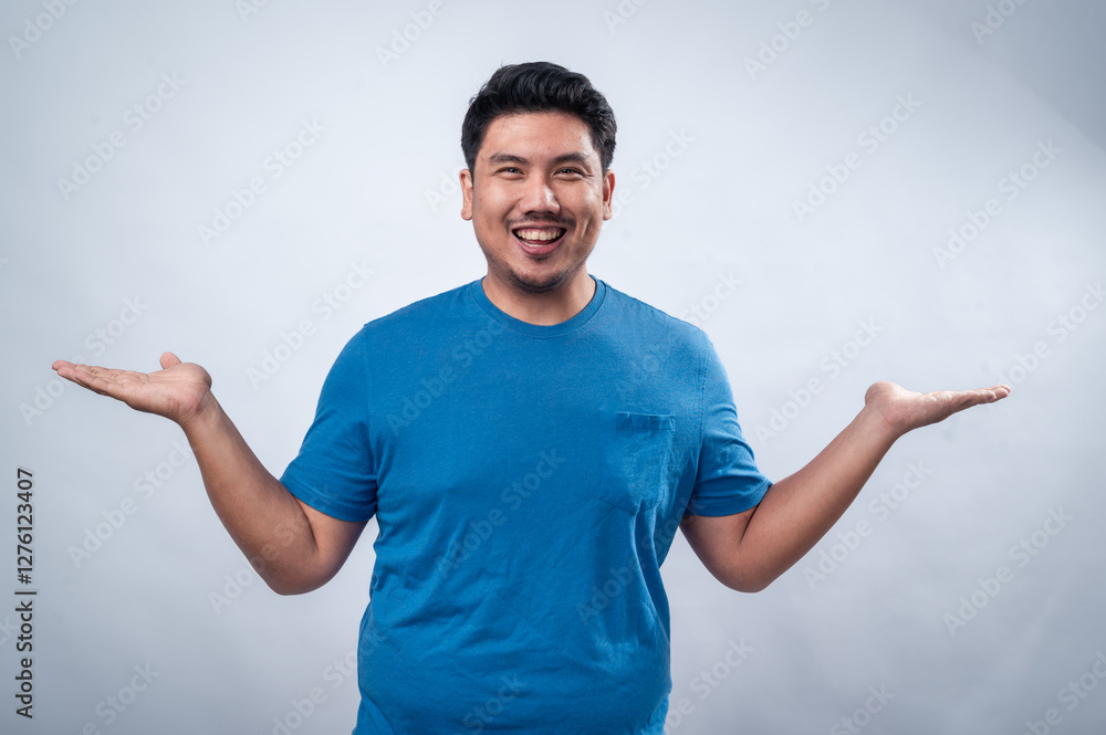 An Asian man in a blue t-shirt stands against a plain white background with both hands raised in a questioning gesture. He smiles warmly, conveying a sense of curiosity, uncertainty, or presentation An Asian man in a blue t-shirt stands against a plain white background with both hands raised in a questioning gesture. He smiles warmly, conveying a sense of curiosity, uncertainty, or presentation