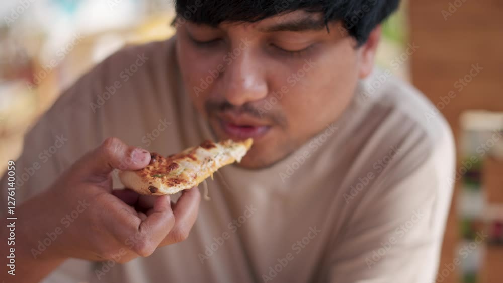 4K Closeup shot of a young Indian man eating pizza while sitting indoors in a café at Manali in India. Man eats junk food whilst sitting in a café. Unhealthy eating background with copy space. 4K Closeup shot of a young Indian man eating pizza while sitting indoors in a café at Manali in India. Man eats junk food whilst sitting in a café. Unhealthy eating background with copy space.