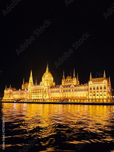 Photography A stunning night view of the illuminated Hungarian Parliament Building in Budapest, Hungary, reflecting beautifully on the Danube River