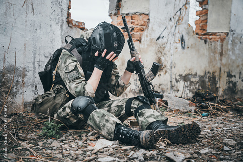 Obraz na płótnie A soldier in uniform with a machine gun sits on the ruins of a destroyed building and covers his face with his hand