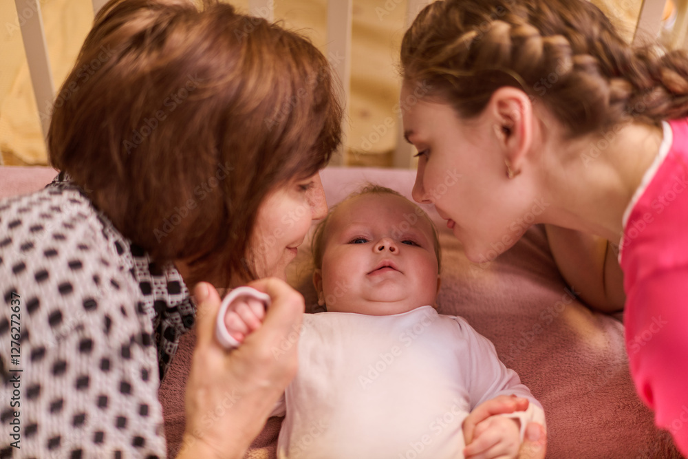Elderly woman with brown hair and young woman with braided hair, both affectionate, lean toward infant inside crib. Soft lighting, warm, intimate atmosphere