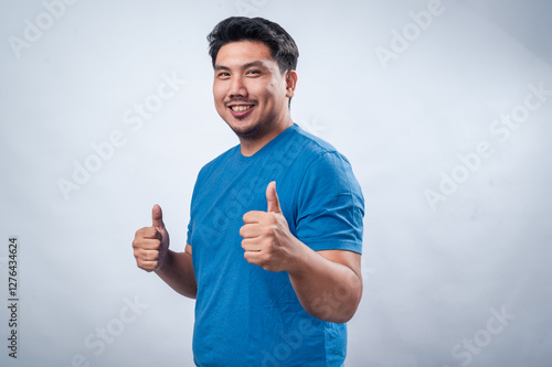 A cheerful Asian man wearing a blue t-shirt gives a thumbs-up gesture while smiling, standing against a clean white background. His positive expression and body language convey confidence and approval photo