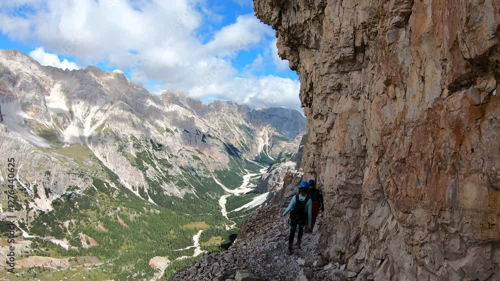 A group of hikers follows a narrow ledge trail with stunning panoramic views of the jagged mountain peaks in the distance. A group of hikers follows a narrow ledge trail with stunning panoramic views of the jagged mountain peaks in the distance.