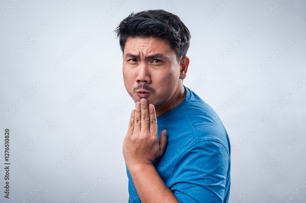 An Asian man wearing a blue t-shirt against a white isolated background is expressing surprise or shock. His mouth is slightly open, and his hand covers it in a playful or astonished manner. An Asian man wearing a blue t-shirt against a white isolated background is expressing surprise or shock. His mouth is slightly open, and his hand covers it in a playful or astonished manner.
