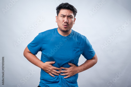 An Asian man in a blue t-shirt against a white isolated background is clutching his stomach with both hands, showing discomfort or pain. His facial expression suggests indigestion, stomachache