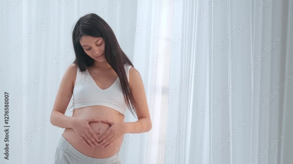 Young happy Asian pregnant woman posing in studio with white curtains in background Young happy Asian pregnant woman posing in studio with white curtains in background