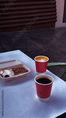 A cup of cappuccino and a cup of coffee with a cake full of chocolate sauce on the white table in the cafe. A picture from afar.