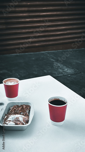 A cup of cappuccino and a cup of coffee with a cake full of chocolate sauce on the white table in the cafe. A picture from afar.