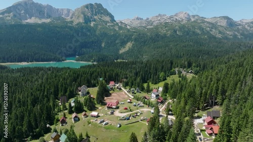 Cinematic drone pullback from a small camp village in a pine forest on the shore of the blue Black Lake, revealing the Durmitor mountain range of the Dinaric Alps in summer