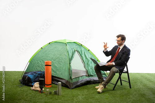 Businessman sitting on chair working on laptop near camping tent, making hand gesture against white studio background. Concept of vacation time, online work, remote business, flexibility. photo