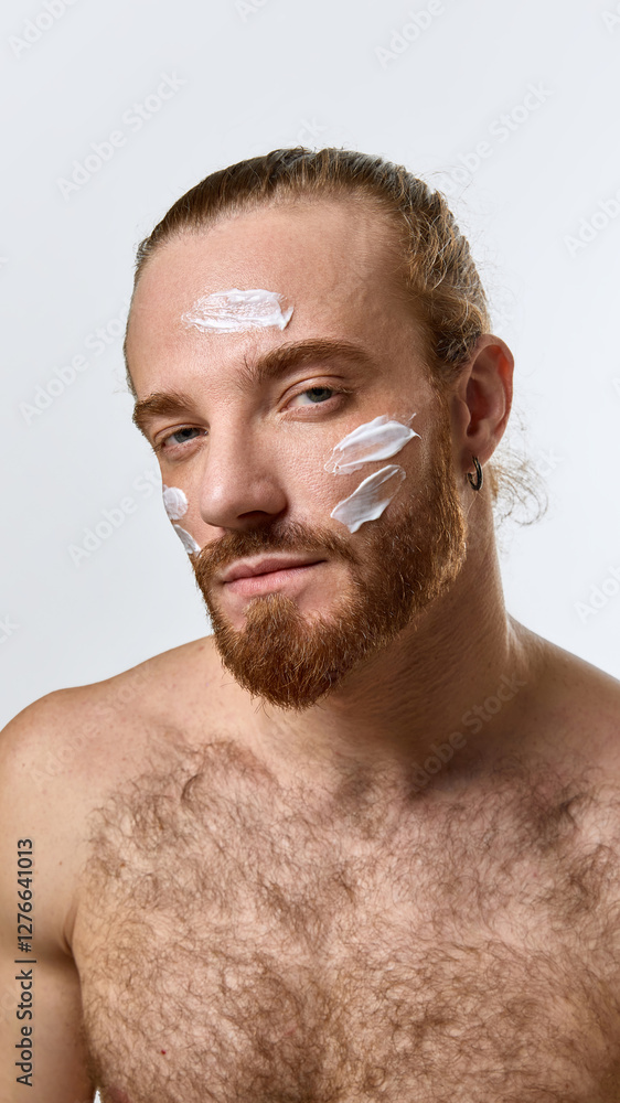 Obraz premium Portrait of handsome bearded shirtless man gazing confidently with white face cream applied across forehead and cheeks, promoting balanced skincare. Concept of male beauty, skin care products