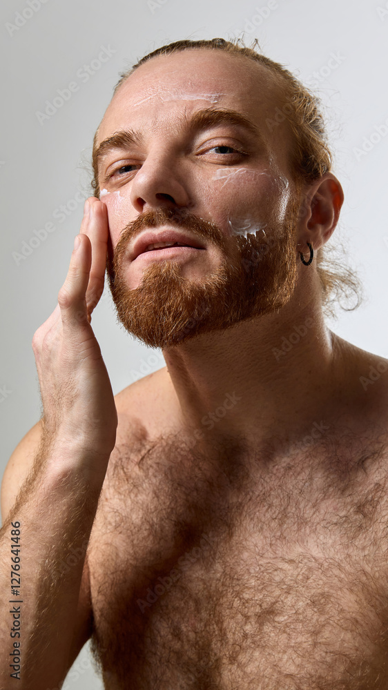 Obraz premium Portrait of handsome man with smooth skin and groomed beard applying cream to face against white studio background. Concept of male beauty, skin care products, healthy skin