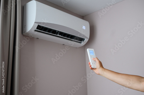 Woman holding air conditioner remote control with display showing 20 degrees Celsius temperature in foreground against air conditioner on wall, sets comfortable cooling temperature on split system