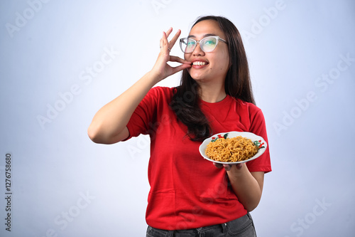 Asian woman with glasses, wearing a red shirt, holding a plate of instant noodles with a satisfied expression, making an 