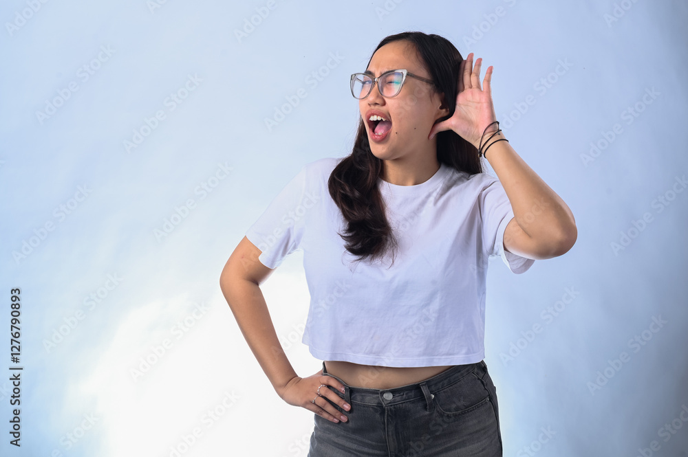 Fototapeta premium Asian woman with long dark hair, wearing glasses, a white crop top, and black jeans, cupping her hand to her ear while smiling. Studio shot with a plain white background, expressing curiosity 