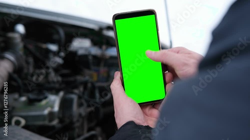 Men's hands are holding a smartphone with a bright green screen against the background of a car engine. Automotive technology. Mobile apps for drivers. Road safety. Using gadgets. Chromakey