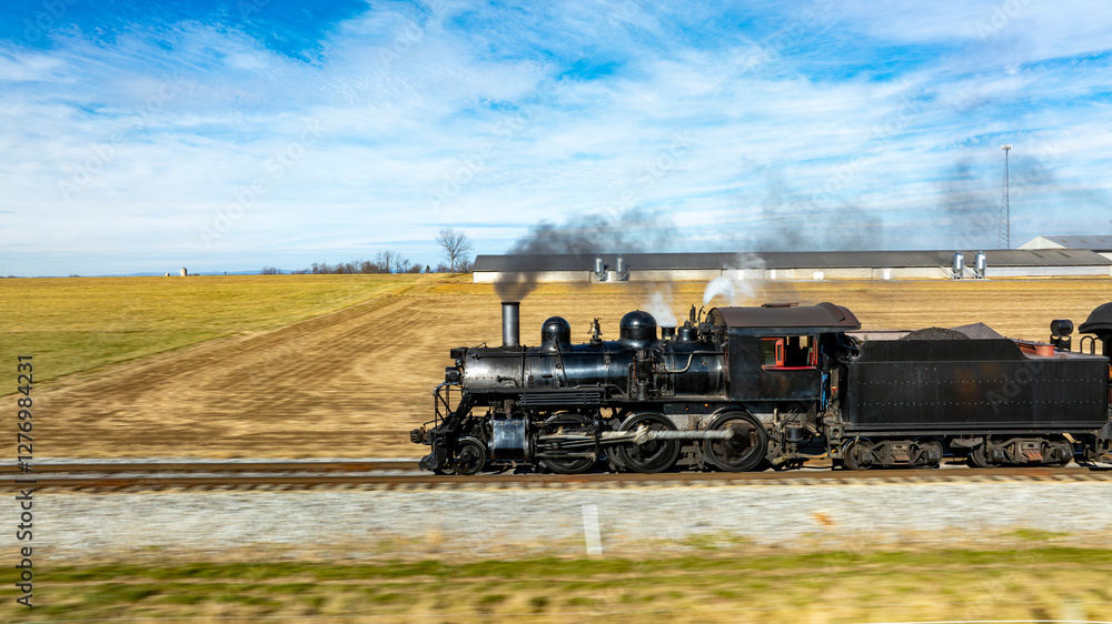 Fototapeta premium A vintage steam locomotive powers along the tracks, sending plumes of smoke into the blue sky. Surrounding fields and farmhouses enhance the picturesque countryside setting.