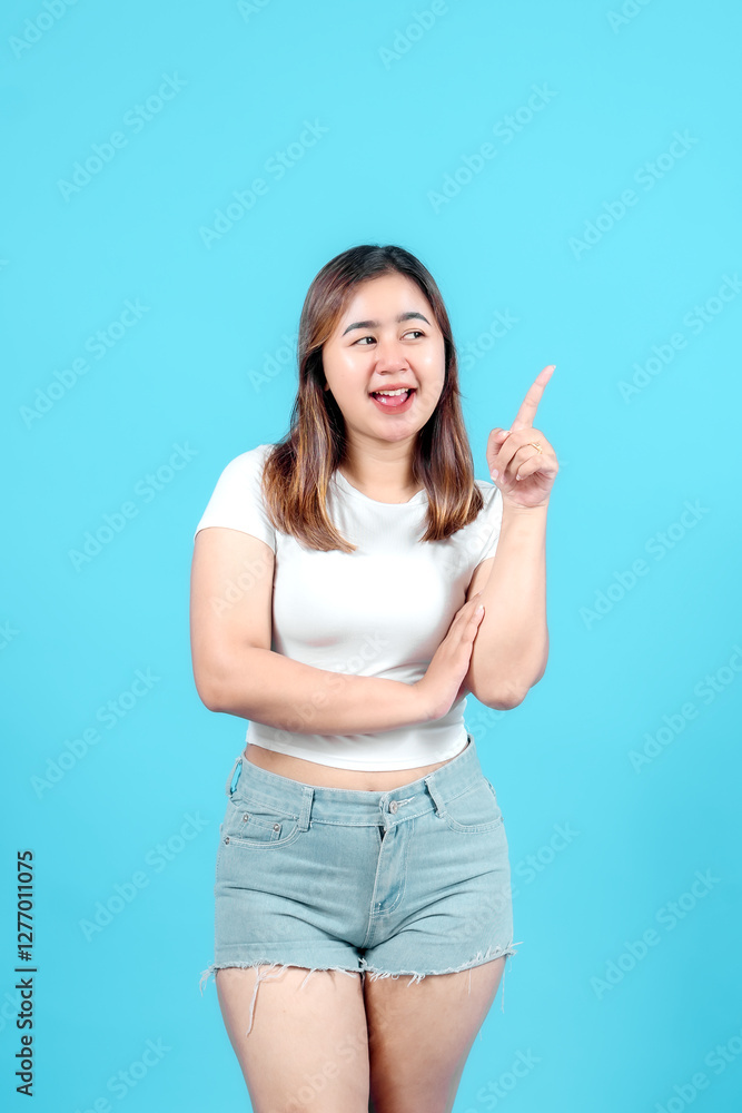 Smiling young woman pointing upwards with one hand, suggesting an idea or direction against a bright blue background. Smiling young woman pointing upwards with one hand, suggesting an idea or direction against a bright blue background.