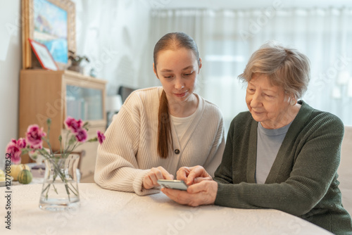 Young woman helping an senior woman with a smartphone at home. Concept of technology, education, and senior digital adaptation. Granddaughter teaches grandmother how to use digital device.