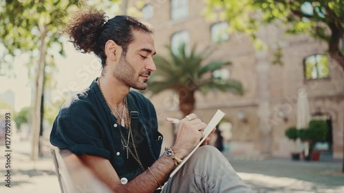 Young italian guy with ponytail and stubble sits on street bench and makes sketches with pen on piece of paper