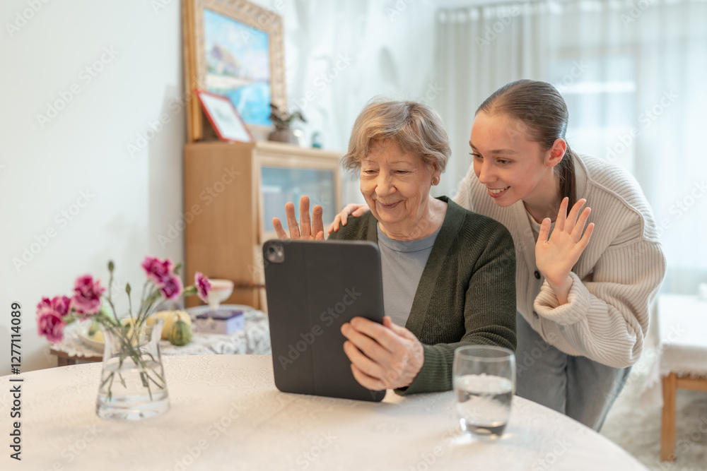 Elderly woman and young woman making a video call with family on a tablet waving to the screen while sitting at home. Concept of technology and family connection. Seniors using digital technology. Elderly woman and young woman making a video call with family on a tablet waving to the screen while sitting at home. Concept of technology and family connection. Seniors using digital technology.