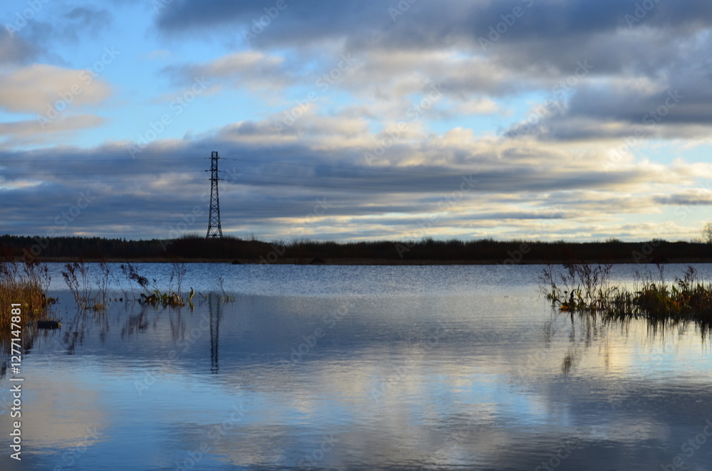Autumn lake. Small waves on the surface of the water, reeds, bushes, trees grow along the banks of the reservoir. The foliage on them has turned yellow. Above, a blue sky with white and gray clouds. Autumn lake. Small waves on the surface of the water, reeds, bushes, trees grow along the banks of the reservoir. The foliage on them has turned yellow. Above, a blue sky with white and gray clouds.