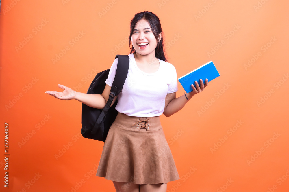 A smiling woman with a backpack and a blue book poses against an orange background ready for study