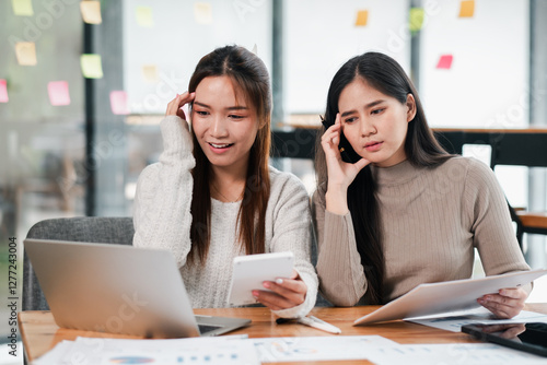 Two young women working together in a modern office, using laptops and documents for a project.