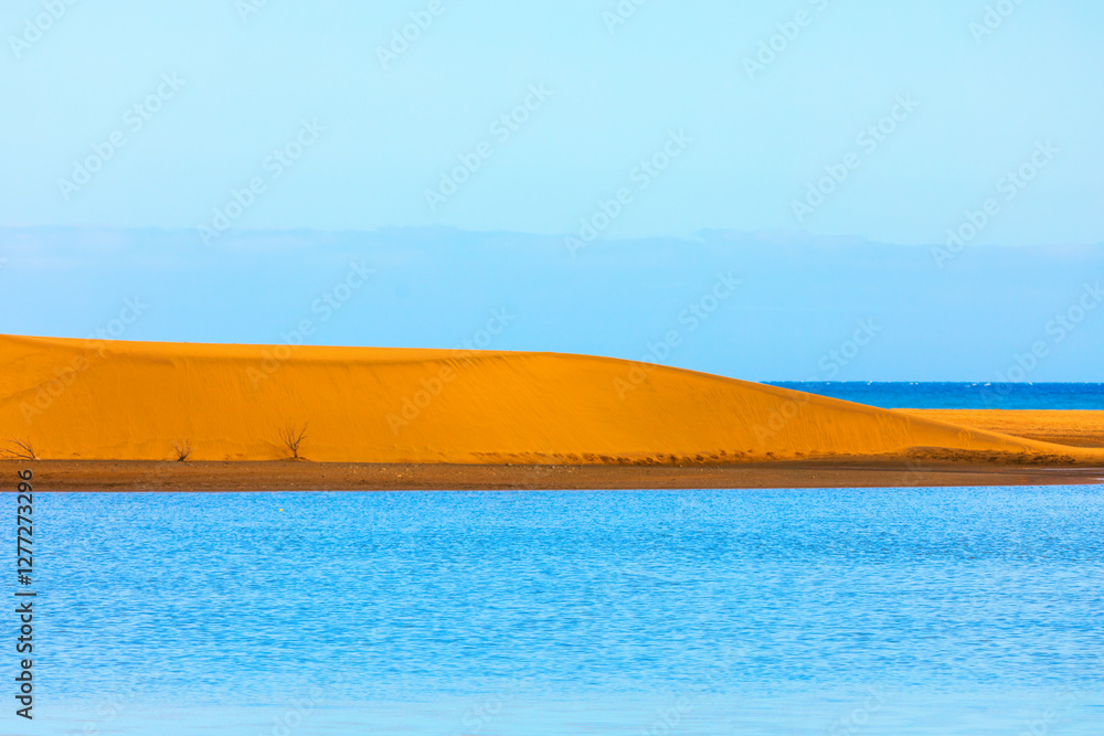 Vibrant landscape of golden sand dune near a blue ocean under clear sky. Contrast between the bright orange sand and the blue water creates a striking and visually appealing scene