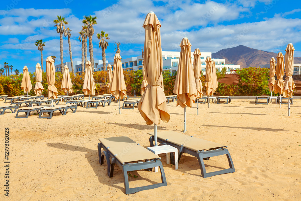 Sandy beach area with multiple beige lounge chairs and closed beige umbrellas arranged in rows. In the background, there are palm trees, a white building, and a mountain under a partly cloudy sky
