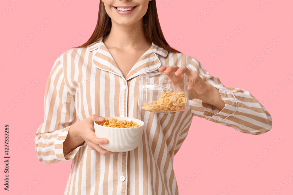Young woman in pajamas pouring tasty cornflakes into bowl on pink background Young woman in pajamas pouring tasty cornflakes into bowl on pink background