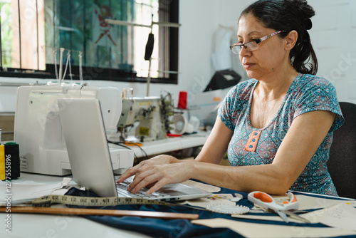 Latin hispanic mid-adult woman using laptop in a sewing workshop, managing fashion business and industry. Technology in small business concept.