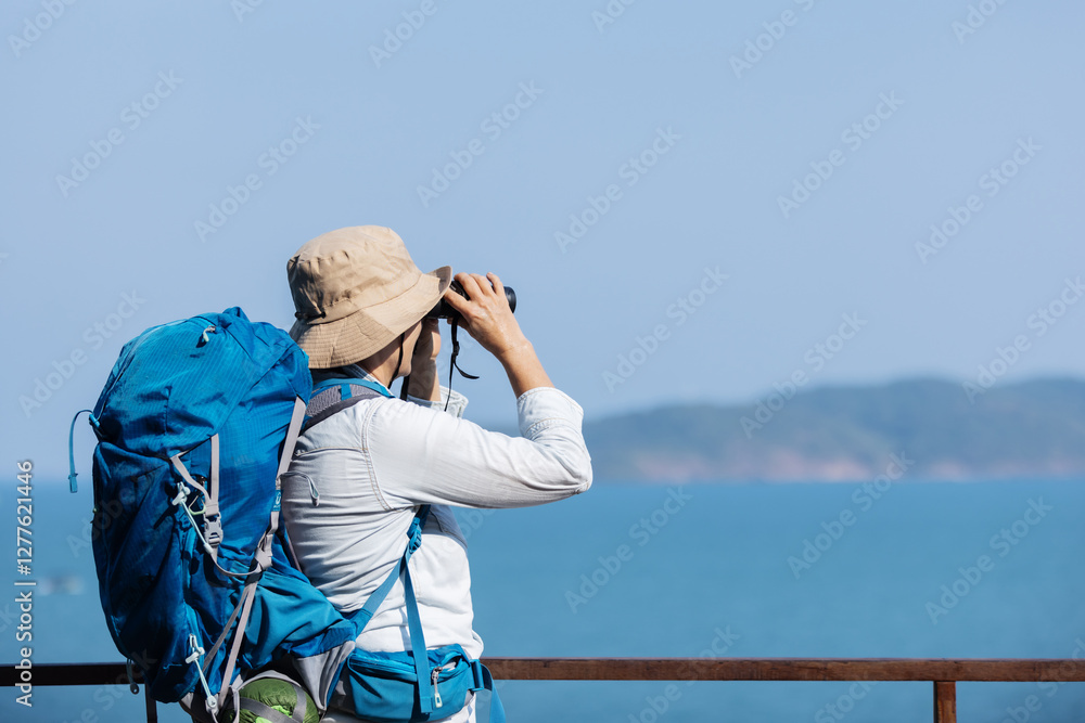 Obraz premium A senior woman with a backpack and hat stands on a viewing deck, using binoculars to observe an island across the ocean. She enjoys the peaceful scenery, fresh air, and the thrill of exploration.