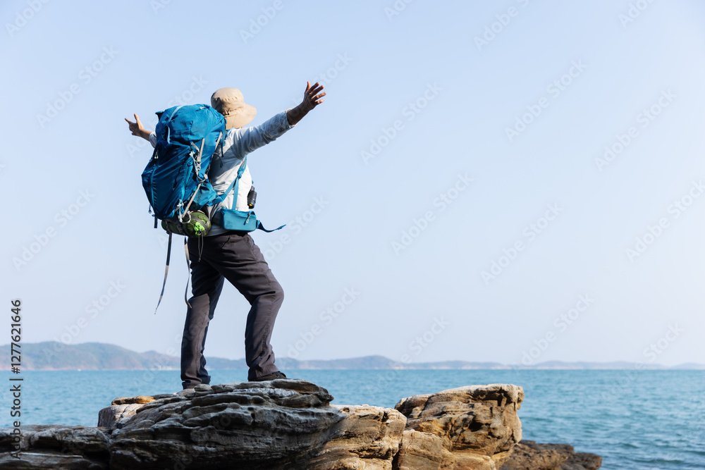 Obraz premium A senior woman stands triumphantly on a rocky shoreline, arms outstretched toward the ocean. Wearing a backpack and hat, she embraces the freedom, adventure, and beauty of the coastal landscape.