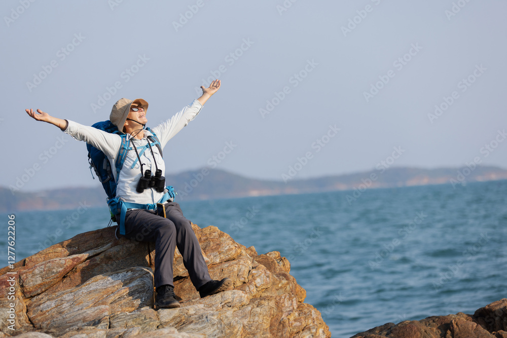 A senior woman sits on a rocky shoreline with arms outstretched, embracing the fresh sea breeze. Wearing a backpack and binoculars, she enjoys the freedom, adventure, and beauty of the ocean landscape A senior woman sits on a rocky shoreline with arms outstretched, embracing the fresh sea breeze. Wearing a backpack and binoculars, she enjoys the freedom, adventure, and beauty of the ocean landscape