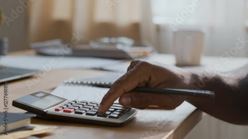Closeup of hands of unrecognizable black man adding up living costs and utility charges on calculator, then writing down sums in notebook, while managing family budget