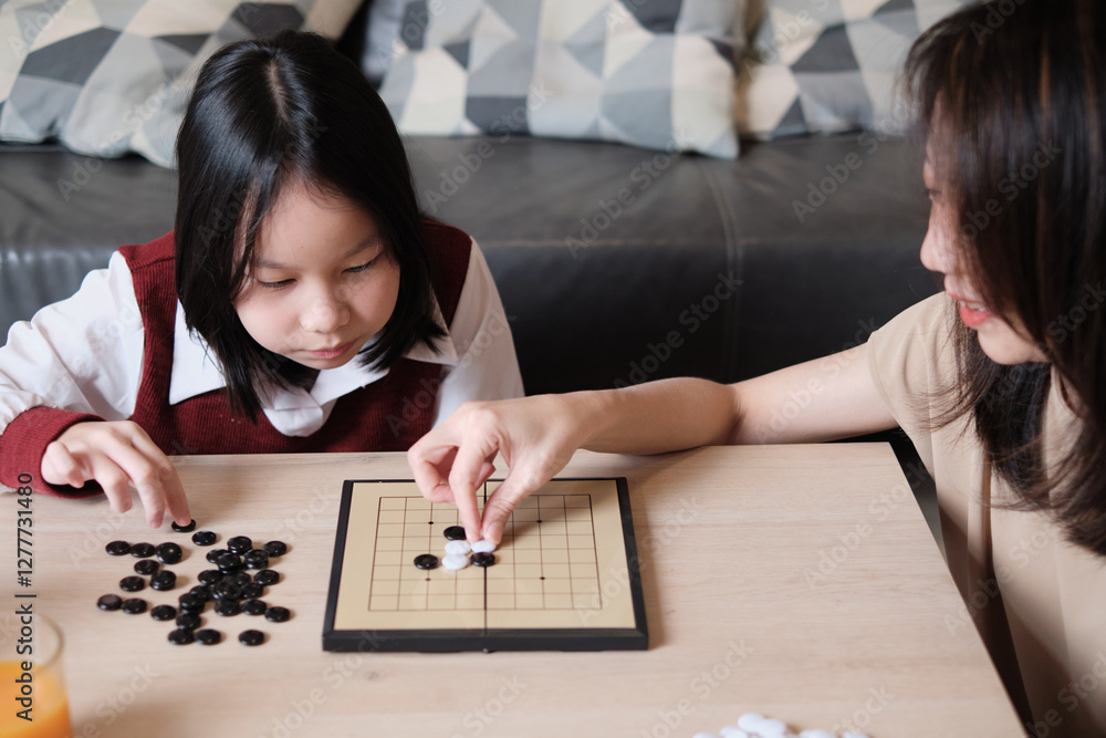 Fototapeta premium Asian family, mum, and kid fun playing Go, checkers board game on table in home living room together with love and cheerful, happy weekend relaxing hobby of child and parent, concentrate and compete.