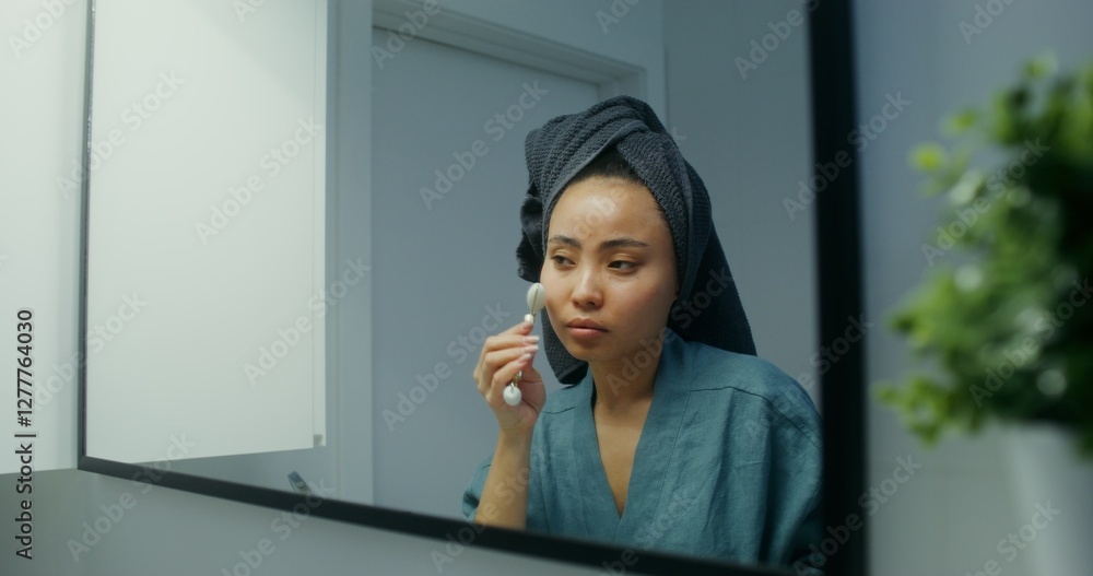 Young asian woman with towel around her head doing facial massage using roller massager while looking at mirror in modern bathroom Young asian woman with towel around her head doing facial massage using roller massager while looking at mirror in modern bathroom