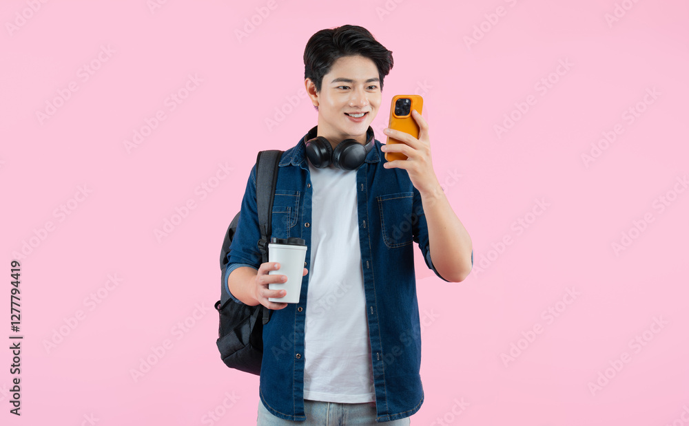 Portrait of Asian male student wearing headphones and backpack , holding cup coffee using phone and posing on pink background Portrait of Asian male student wearing headphones and backpack , holding cup coffee using phone and posing on pink background
