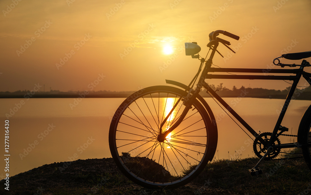 Fototapeta premium Silhouete of old rusty retro style bicycle stands in a meadow near a lake at sunset.