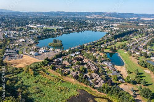Aerial drone view of Staats Lake in Keizer, Oregon, near Salem, featuring a residential neighborhood, a golf course, and lush green landscapes under a clear blue sky
